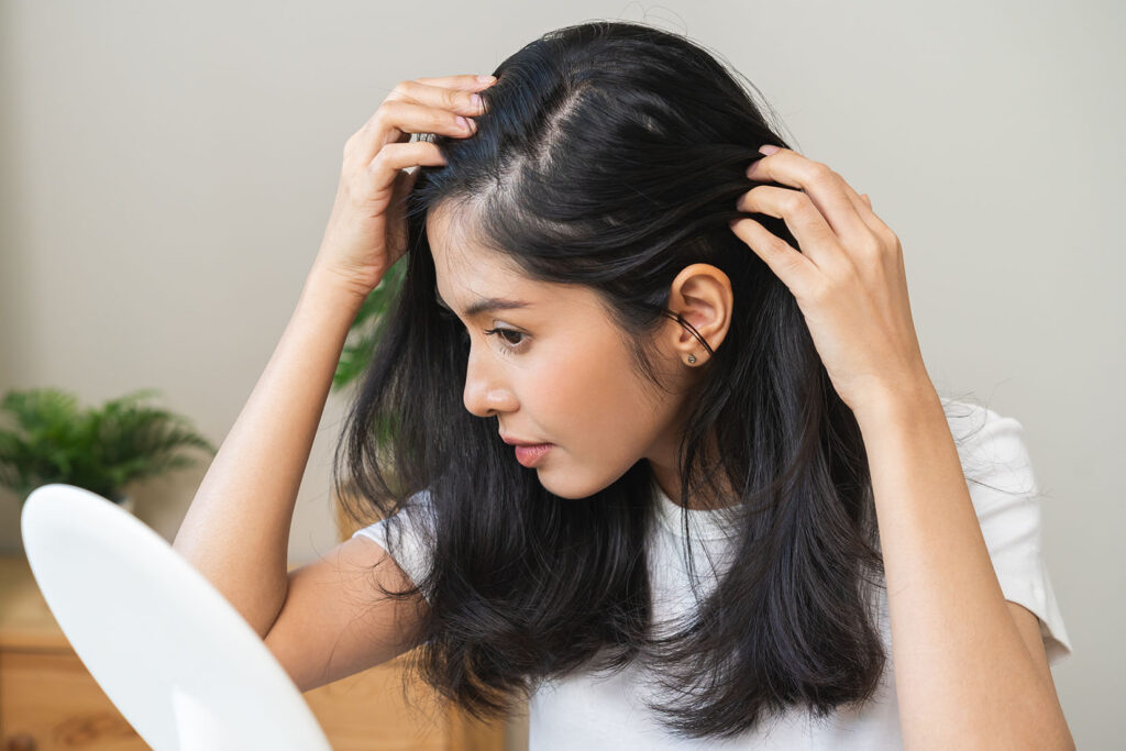 woman with long dark hair parting her hair and looking at her scalp in a mirror