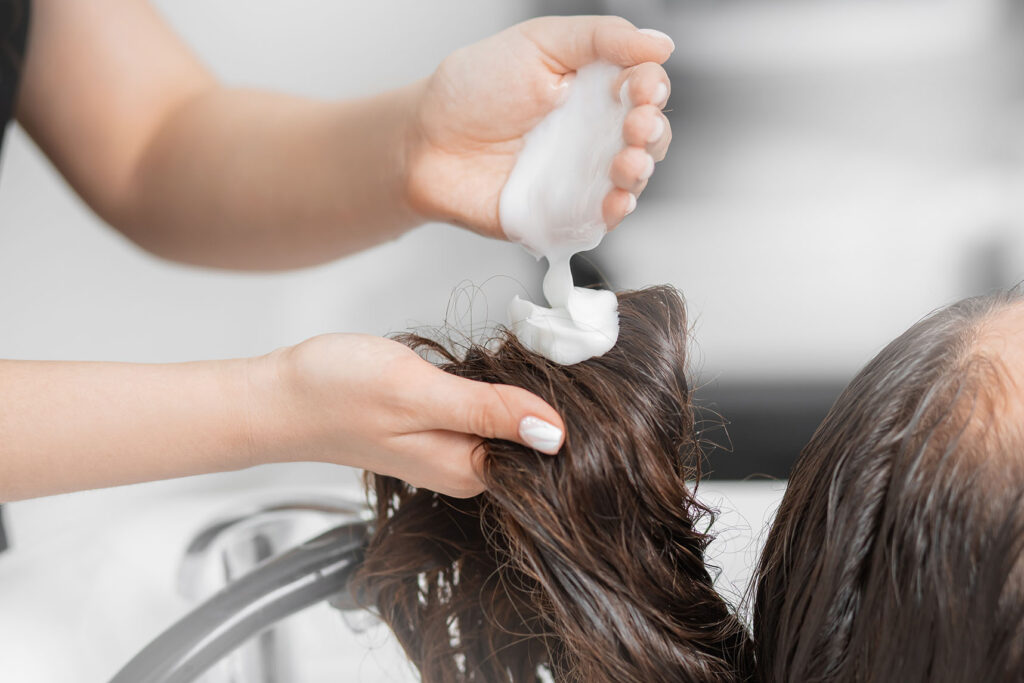 hands applying a thick white cream onto wet hair over a salon basin