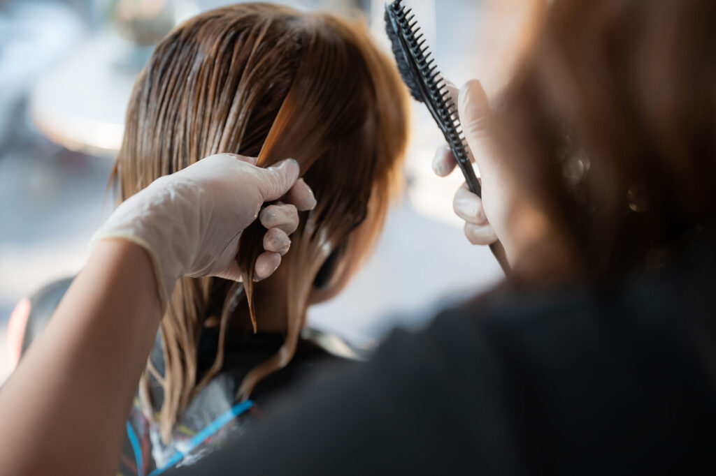 Close-up of a stylist applying keratin solution to wet hair using gloved hands and a comb.